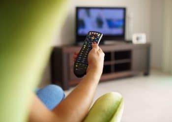 Over the shoulder view of girl sitting on sofa holding tv remote and surfing programs on television