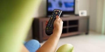 Over the shoulder view of girl sitting on sofa holding tv remote and surfing programs on television