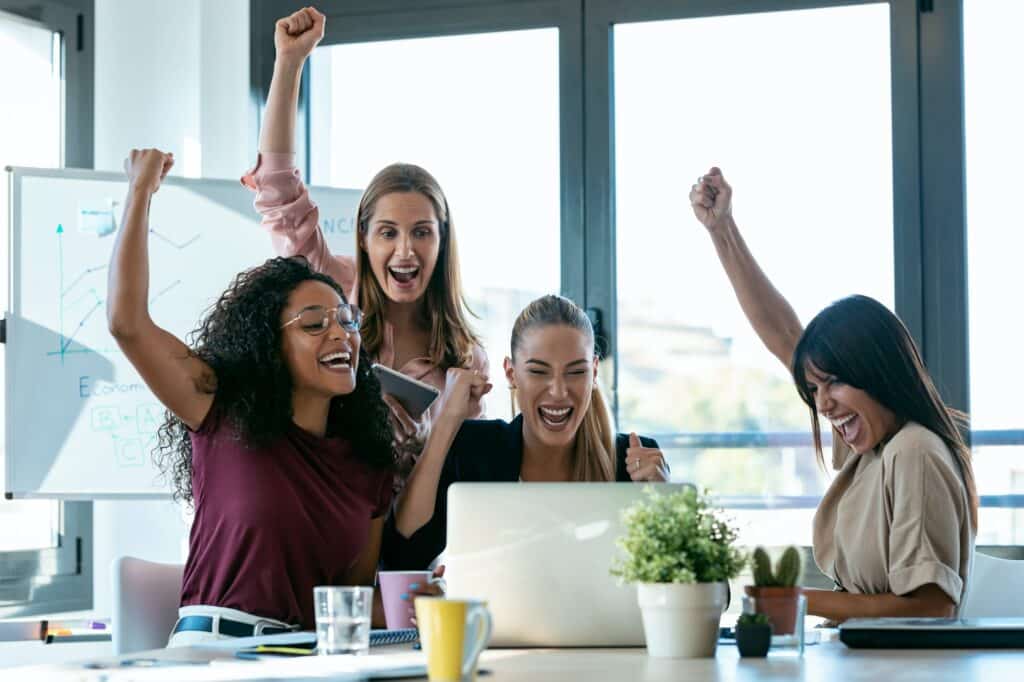 Four happy smart business women work with laptops while celebrating a victory in a coworking space
