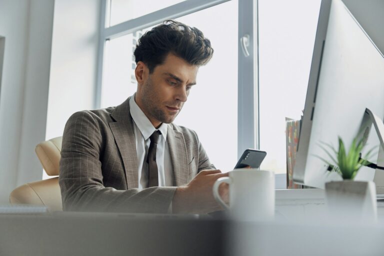Concentrated young man using smart phone while sitting at his working place in office